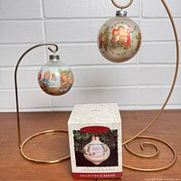 Two vintage Betsey Clark glass Christmas ornaments displayed on gold metal stands with a white wall background, one ornament hanging in original packaging box.