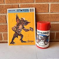 Photo of framed Gremlins Stripe puzzle and vintage Gremlins thermos side by side against brick background