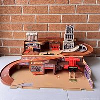 Front view of the multi-level brown plastic Hot Wheels city playset with ramps, parking elevator, garage, and building facades against a brick wall.