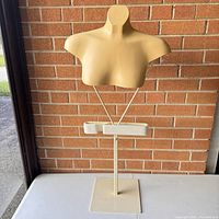 Full view of beige vintage female mannequin bust mounted on adjustable metal rod stand and square metal base against a brick wall background.