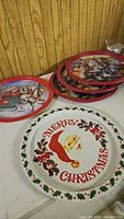 Photo showing five round metal trays stacked on a table against a wood paneled wall. One tray in the front has a 'Merry Christmas' Santa face design with holly border. Another visible tray shows Santa riding a reindeer sleigh.