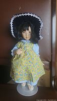 Front view of vintage 14 inch Cottage Collectibles doll by Ganz wearing blue and white striped blouse, yellow floral dress apron, black bonnet with white lace, standing on white plastic stand.