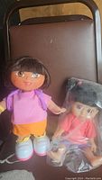 Photo shows two battery operated dolls placed on a brown chair. Dora doll wearing pink shirt and orange shorts, Boo doll in pink outfit in plastic bag.