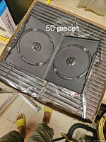 Open view of a black plastic DVD jewel case showing disc hub and inner structure. Stack of 50 cases still sealed in plastic packaging visible underneath.