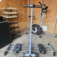 Full view of the silver Leg Magic exercise machine with black foot platforms and stationary bike in background