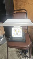 Manual baby nursery scale with white weighing tray, dial with baby-themed markers, positioned on a brown chair indoors.