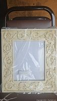 Picture frame with a detailed cream-colored floral and swirl pattern, sealed in plastic wrap.