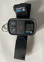 Top view of a black GoPro remote control with an LCD screen and buttons, attached to a black adjustable strap with a metal ring.
