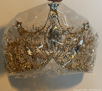 Front view of the heavy tiara crown, showing intricate metalwork with round and teardrop clear gemstones, wrapped in plastic.