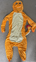 Full view of the orange and beige Dragonite onesie laid flat on a carpeted floor showing front closure buttons, hood details including face, eyes, and teeth, and tail with red tip.