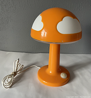 Front view of the IKEA Skojig orange mushroom lamp showing the dome shade with white clouds, orange stem and round base with white push-button switch and attached white cord.