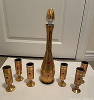 Gold-colored wine decanter with stopper and six matching gold goblets featuring red jewel and ornate designs arranged on floor against white door backdrop.
