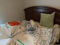 Queen size white duvets with green stickers, orange quilted throw, bolster pillows, beige patterned pillow shams, blue packaged throw on bed with wooden headboard.