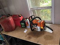 Full view of chainsaw, two red fuel jugs, MotorMaster chain oil and Stihl engine oil on workbench.