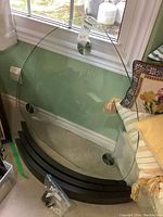 Clear glass triangular coffee table top with rounded edges resting against a wall near a window, dark wooden curved base at the bottom, hardware bag visible on floor.