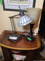 Pair of two different table lamps on a wooden side table. One lamp has a flexible arm and a white segmented glass shade. The other has a frosted textured glass shade on a wooden base.
