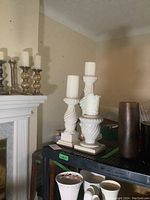 Photo showing the white porcelain pillar candle sticks of varying heights arranged on a black surface near a fireplace mantel, with accompanying white and textured candles on top.