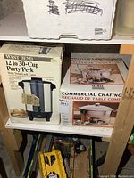 West Bend 12 to 30-Cup Party Perk coffee maker and commercial chafing dish in their original boxes, shown on wooden shelving.