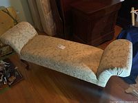 Full view of the gold-colored fabric chaise lounge with curved padded ends and decorative wooden legs on a hardwood floor.