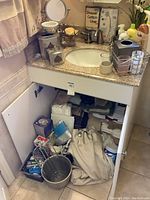 Under-sink cabinet open with various toiletries, wipes, containers, metal basket, and textiles packed inside.