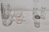 Full set of assorted glass kitchenware including milk jars, measuring cups, sugar shaker, and pitchers on white surface.