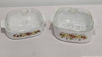 Photo showing both vintage Pyrex casserole dishes side by side on a white surface, each with glass lids and colorful harvest pattern on the side.