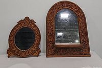 Two hand crafted wooden mirrors side by side; left is an oval mirror with floral carvings, right is an arched mirror with floral carvings and a shelf