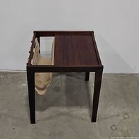 Full view of mid-century wooden newspaper table showing dark wood frame, wooden top, and canvas sling holder underneath from side angle.