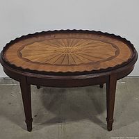 Front and side view of vintage oval wooden table showing carved scalloped edge and two-toned inlay sunburst pattern on tabletop.