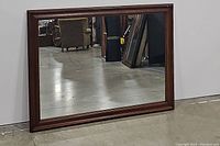 Front view of the large rectangular wooden framed mirror leaning against a wall on a concrete floor.