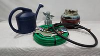Photo showing all items in lot including watering can, solar angel figurine, ceramic water fountain, green garden hose, and black extension hose