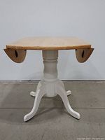 Front view of the drop leaf dining table showing the natural wood top with two drop leaves and white painted pedestal base with four legs.