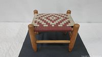 Woven wooden foot stool showing angled side view highlighting the rounded legs and the red and beige woven pattern on the top.