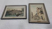 Two framed prints by Paul Lambert of Parisian street scenes side by side on a white quilted surface.