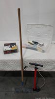 Photo of metal garden rake with wooden handle, red air pump, and a transparent plastic bin containing other tools