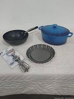 Photo showing all items in lot: two non-stick wok-style frying pans, Le Creuset cobalt blue Dutch oven with lid, non-stick pie tin, and rotary hand beater in package.