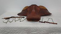 Full view of the brown leather saddle laid flat with four metal spurs in front with leather straps and two pairs of wooden and metal boot hooks on the right side.
