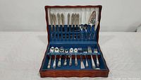 Wooden case open showing two sets of flatware displayed on blue felt lining. Upper tier contains silver flatware with ornate handles, including serving utensils. Lower tier contains additional cutlery with simpler design.
