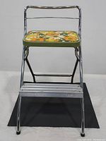 Front view of vintage folding step stool showing floral patterned top and chrome metal frame with footrest.