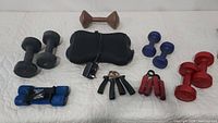 Full view of the collection showing all dumbbells in black, blue, red, and brown colors, three hand grip strengtheners, and the black electric massage pad with power cord.
