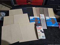 Six beige, black, and brown sanding sheets, two packaged Sears KARBO-GRIT tungsten carbide sheets, a packaged sanding belt, and several loose sheets on a black surface.