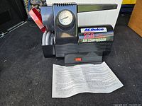 Air compressor front view showing AC Delco logo, pressure gauge, power switch, attached hose, and manual.