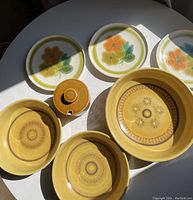 Photo of three white round plates with floral design and four yellow bowls with circular brown patterns, plus a small covered mustard pot