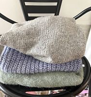 Three folded sweaters stacked on black chair: light gray on top, blue chunky knit in middle, light green at bottom