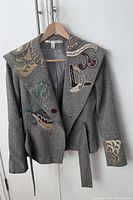 Full front view of grey wool blend jacket featuring multi-color embroidery on collar, front and sleeves. Hanging on wooden hanger.