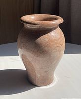 Heavy marble vase with pinkish undertone standing on a white surface with sunlight casting shadows.