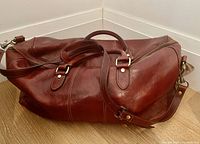Side angle view of reddish brown leather bag with handles and zipper closure lying on wood floor.