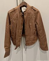 Front view of brown vegan leather jacket with snap-button closure, long sleeves, and collar, hanging on a wooden hanger.