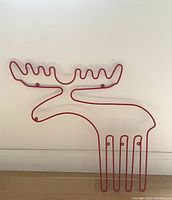 Red wire moose-shaped metal coat rack resting against white wall on hardwood floor, showing full profile with broad antlers and legs.