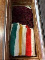 View inside the wooden chest showing the folded wool blanket with green, yellow, and red stripes and partially visible burgundy knitted throw.